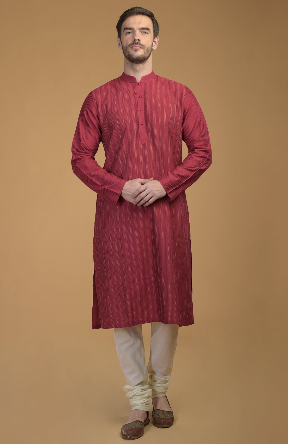 A man stands wearing a wine-colored kurta with pintuck and kantha details and ivory churidars.