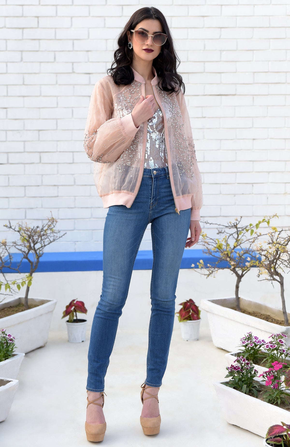 A woman wears a sheer peach bomber jacket with crystal flower embroidery over a sequined top and jeans.