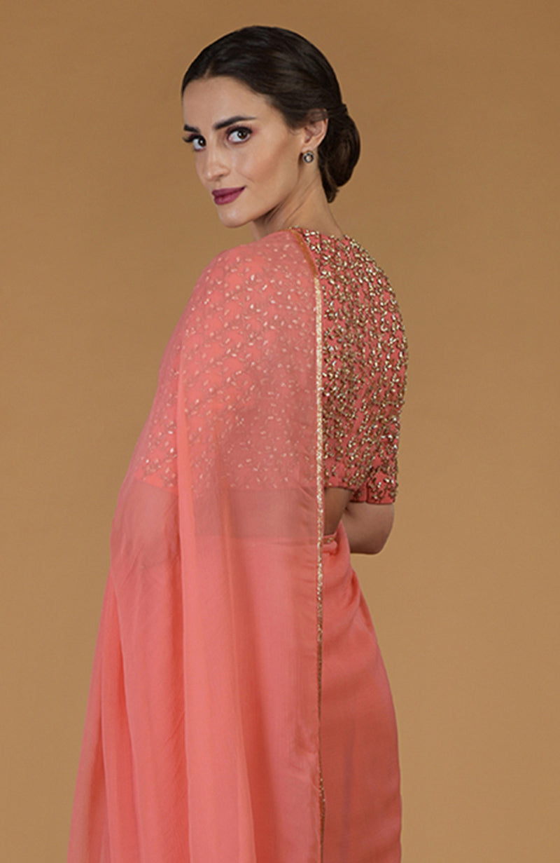 A woman in a salmon pink saree with a heavily embroidered blouse looks over her shoulder.
