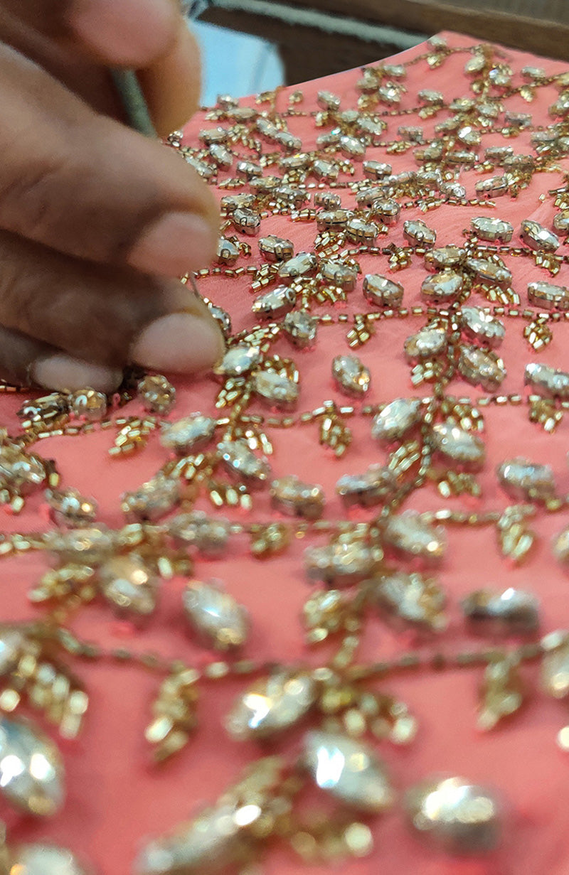 A close-up of a hand embroidering a salmon blouse with gold beads and clear crystals.